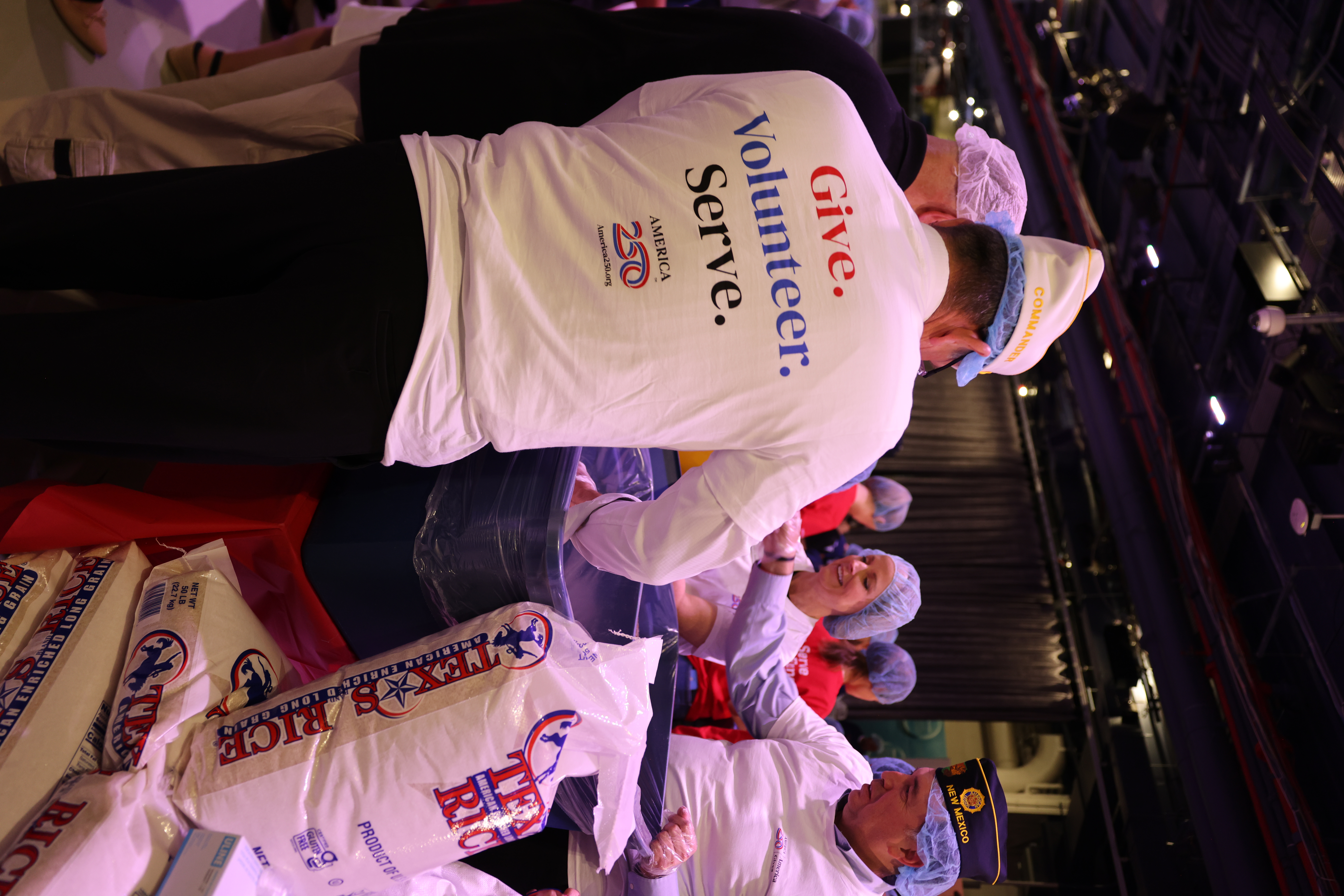 Veterans and volunteers pack food at a service event, wearing shirts that say “Give. Volunteer. Serve.”